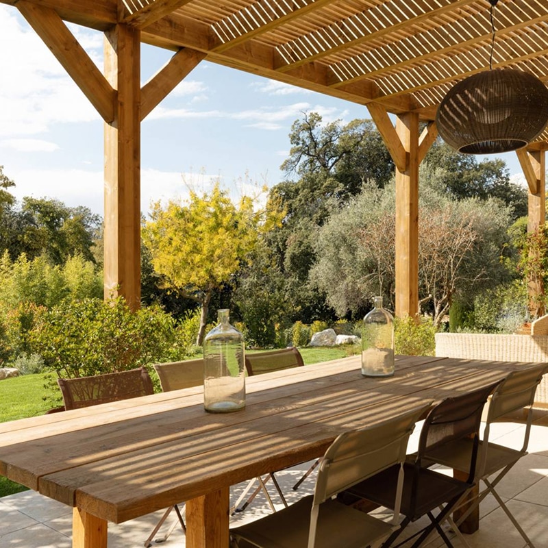Pergola en bois sur terrasse ensoleillée avec longue table et chaises, entourée d'un jardin luxuriant. Deux carafes vides sur la table.