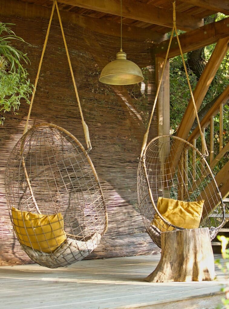 Deux fauteuils suspendus en métal ajouré avec coussins jaunes sur une terrasse en bois, sous une lampe industrielle.