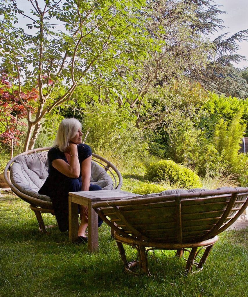 Femme aux cheveux blonds assise dans un fauteuil rond en rotin dans un jardin verdoyant, regardant au loin.
