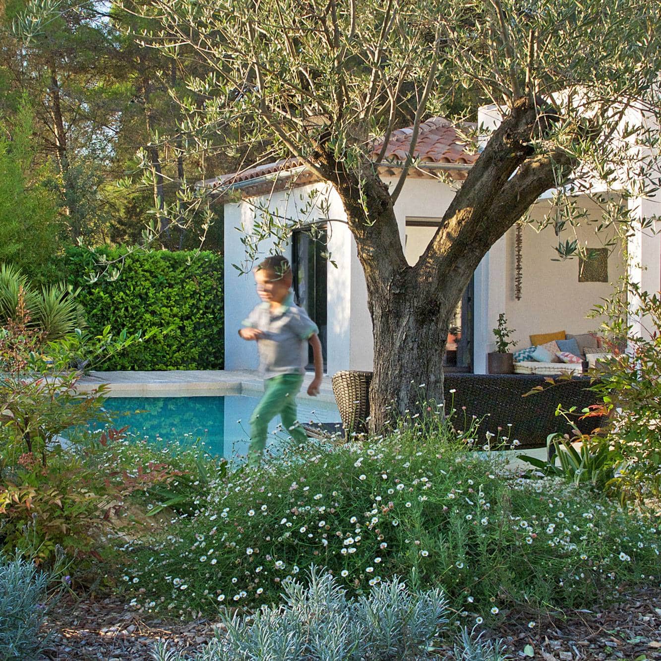 Jeune garçon en mouvement près d'une piscine, devant un olivier et une maison de style méditerranéen avec terrasse fleurie.