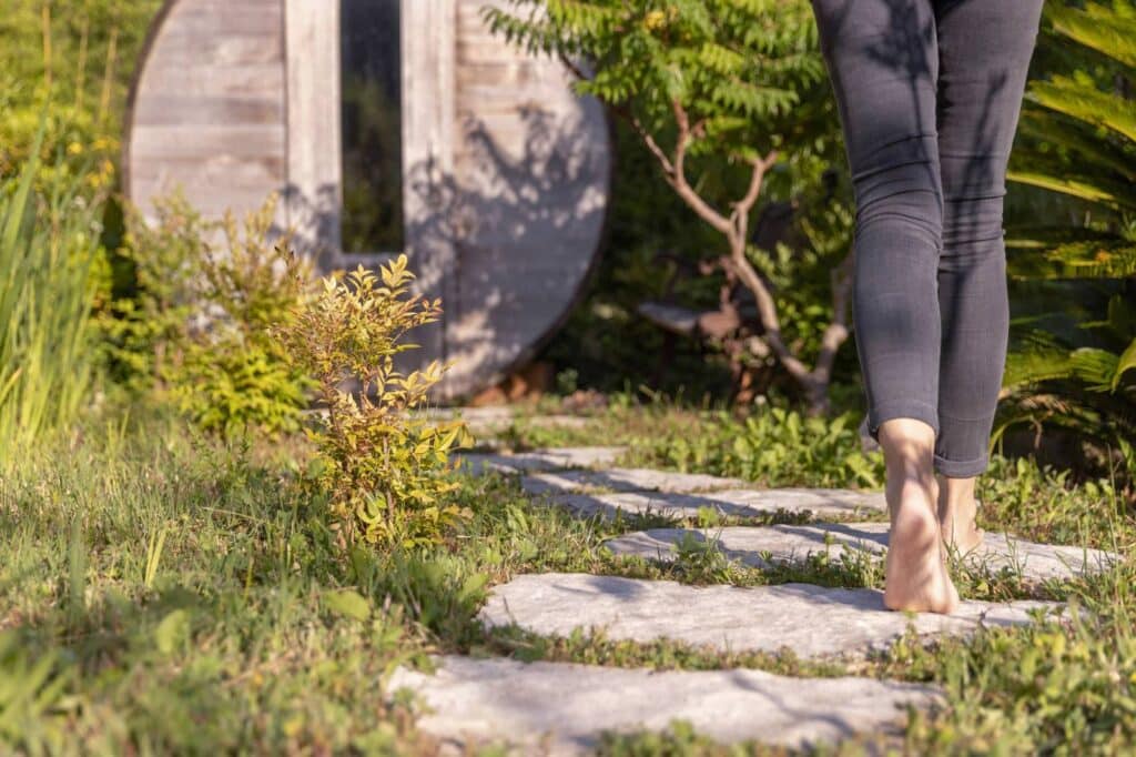 Jambes d'une personne pieds nus marchant sur un chemin de dalles de pierre à travers l'herbe vers un sauna en bois extérieur.
