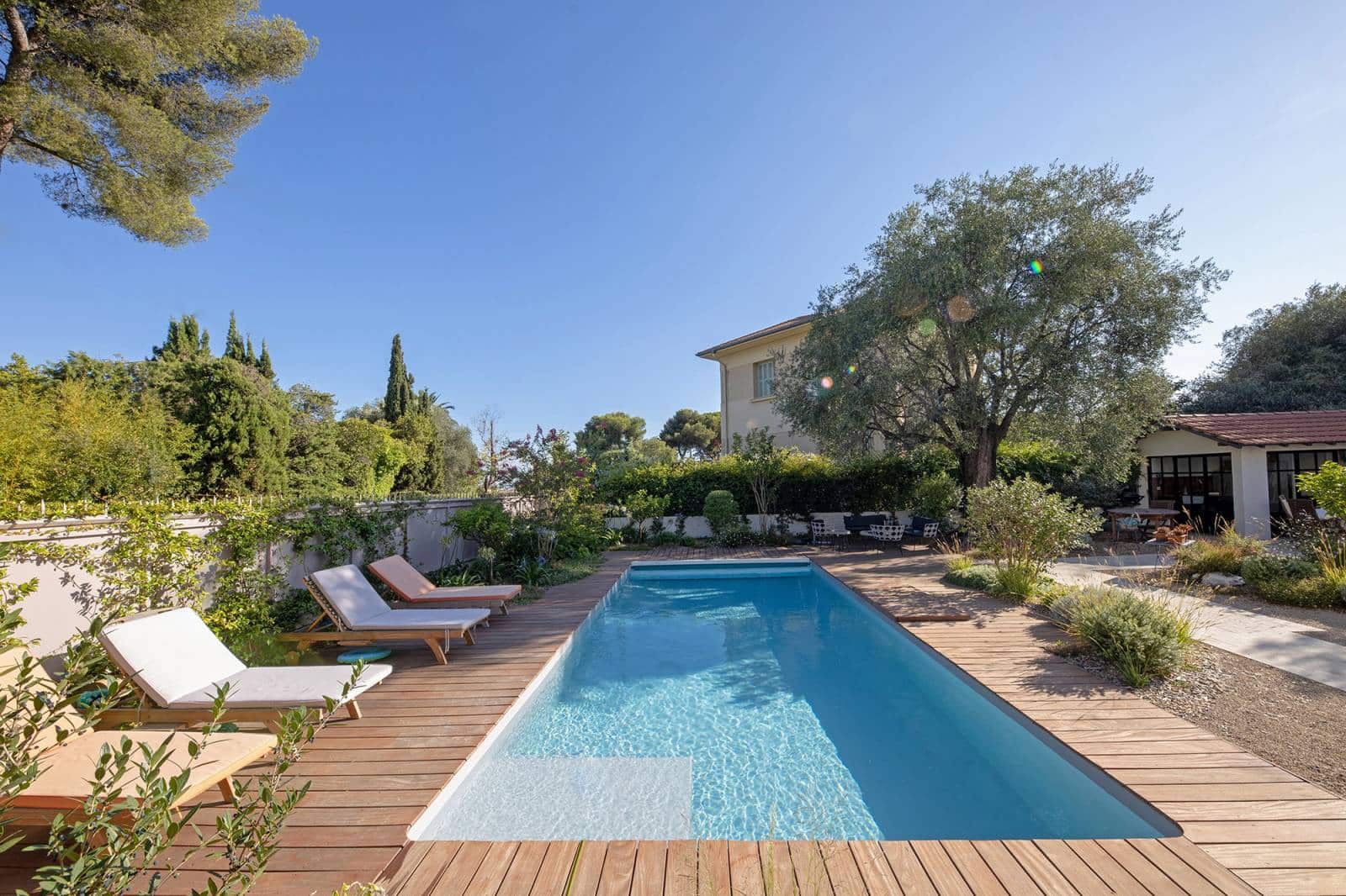 Piscine rectangulaire bordée de bois clair, avec chaises longues, entourée d'une végétation luxuriante et d'une maison provençale.