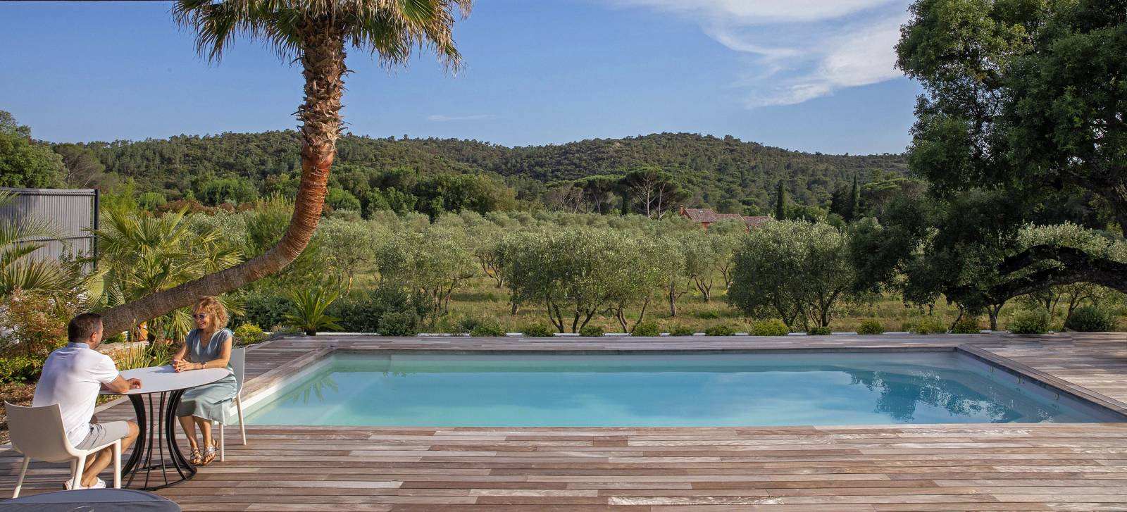Couple discutant au bord d'une piscine moderne avec terrasse en bois, vue sur oliveraie et collines boisées.