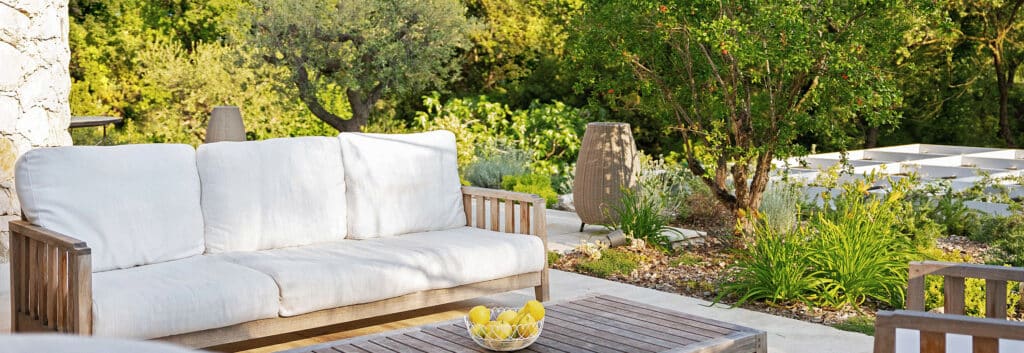 Ambiance détente sur un canapé en bois et coussins blancs. Terrasse donnant sur un jardin luxuriant et un arbre fruitier.