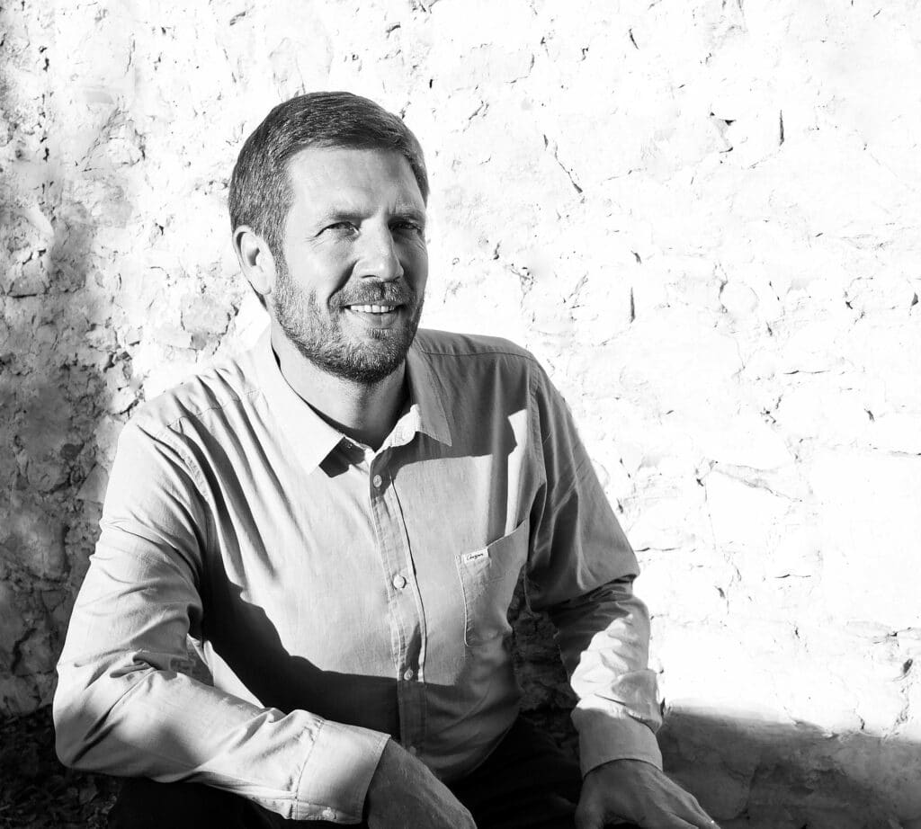 Homme souriant en noir et blanc, portrait extérieur Portrait en noir et blanc d'un homme souriant, barbe soignée, en chemise claire, assis contre un mur de pierre brut en plein soleil.